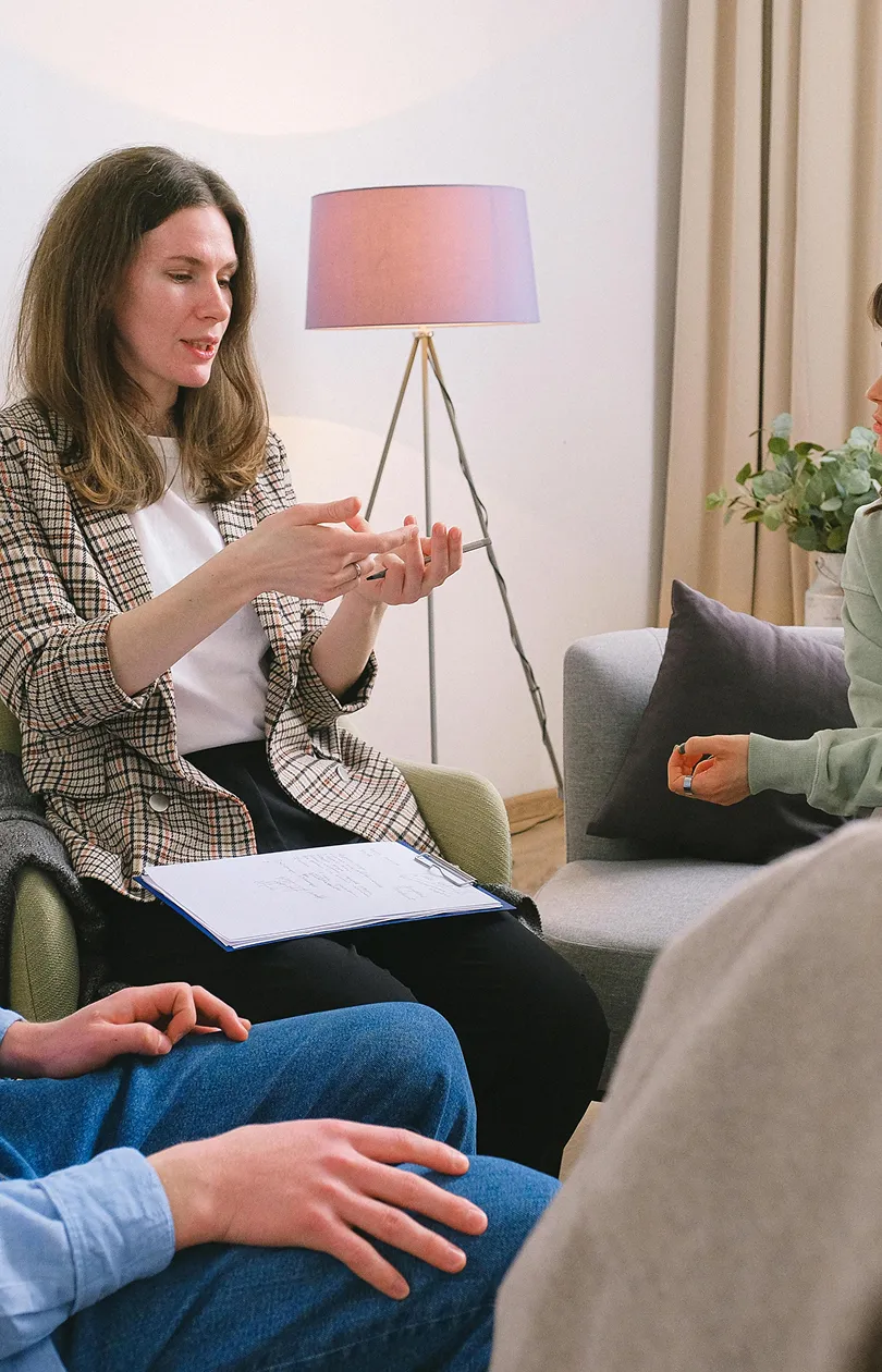 A woman in a plaid blazer holding a pen and clipboard, engaged in conversation with two other people seated nearby, in a cozy room with a floor lamp and plants.