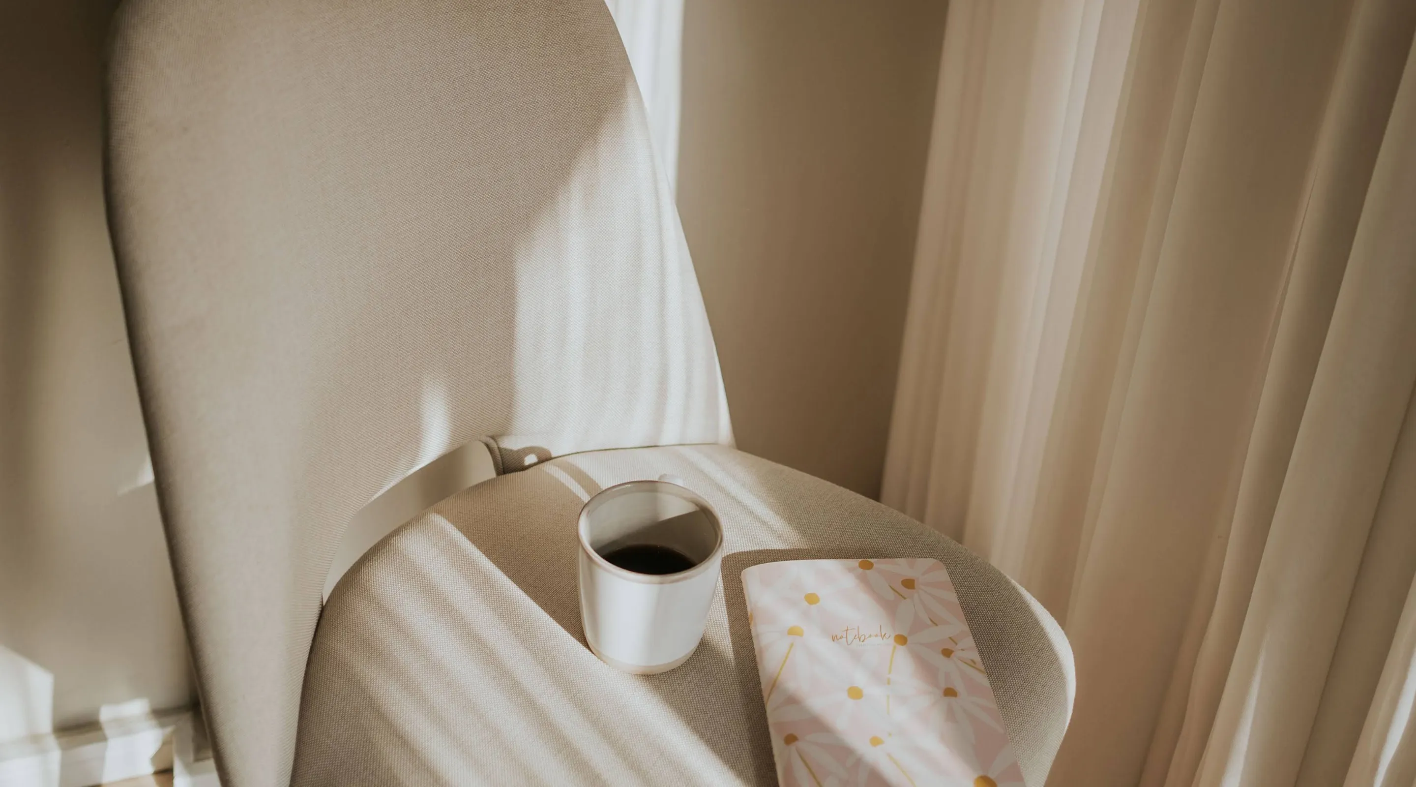 Beige chair with stripes of light and shadow, holding a cup of coffee and a floral notebook, next to beige curtains.