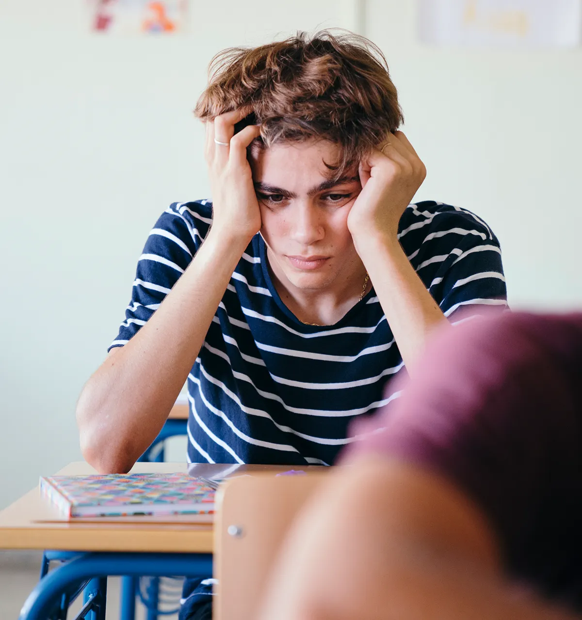 Anxious teenage boy sitting at a school desk with his head in his hands.
