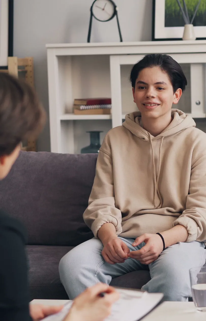 Smiling teenage boy sitting on a couch during a therapy session with a clinician taking notes.