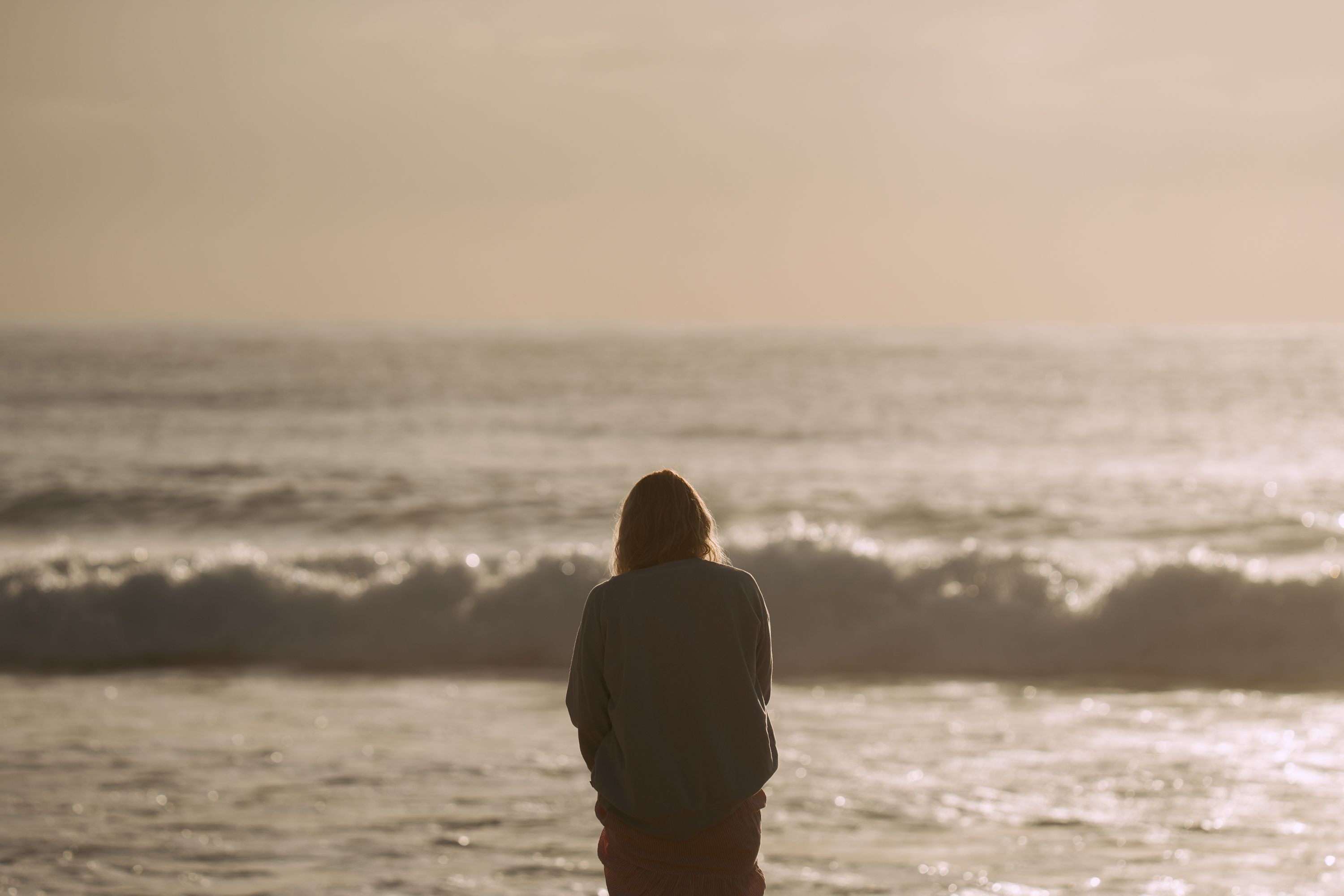 Person standing on a beach facing the sea during sunset with waves gently rolling in.