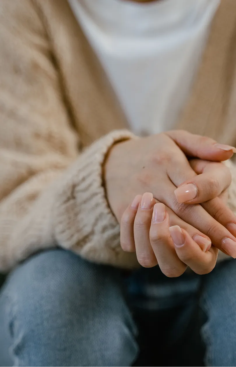 Close-up of a person sitting with hands gently clasped, wearing a beige knitted sweater and blue jeans.