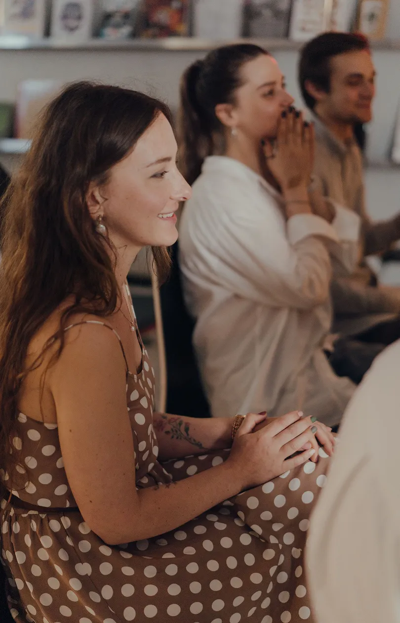 Three people sitting in a row indoors; a smiling woman in a brown polka dot dress is in the foreground, with a woman in white and a man in the background, both slightly blurred.