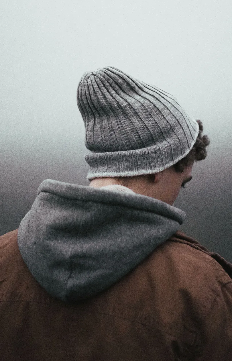 Person seen from behind wearing a gray knit beanie, a gray hood, and a brown jacket against a foggy background.