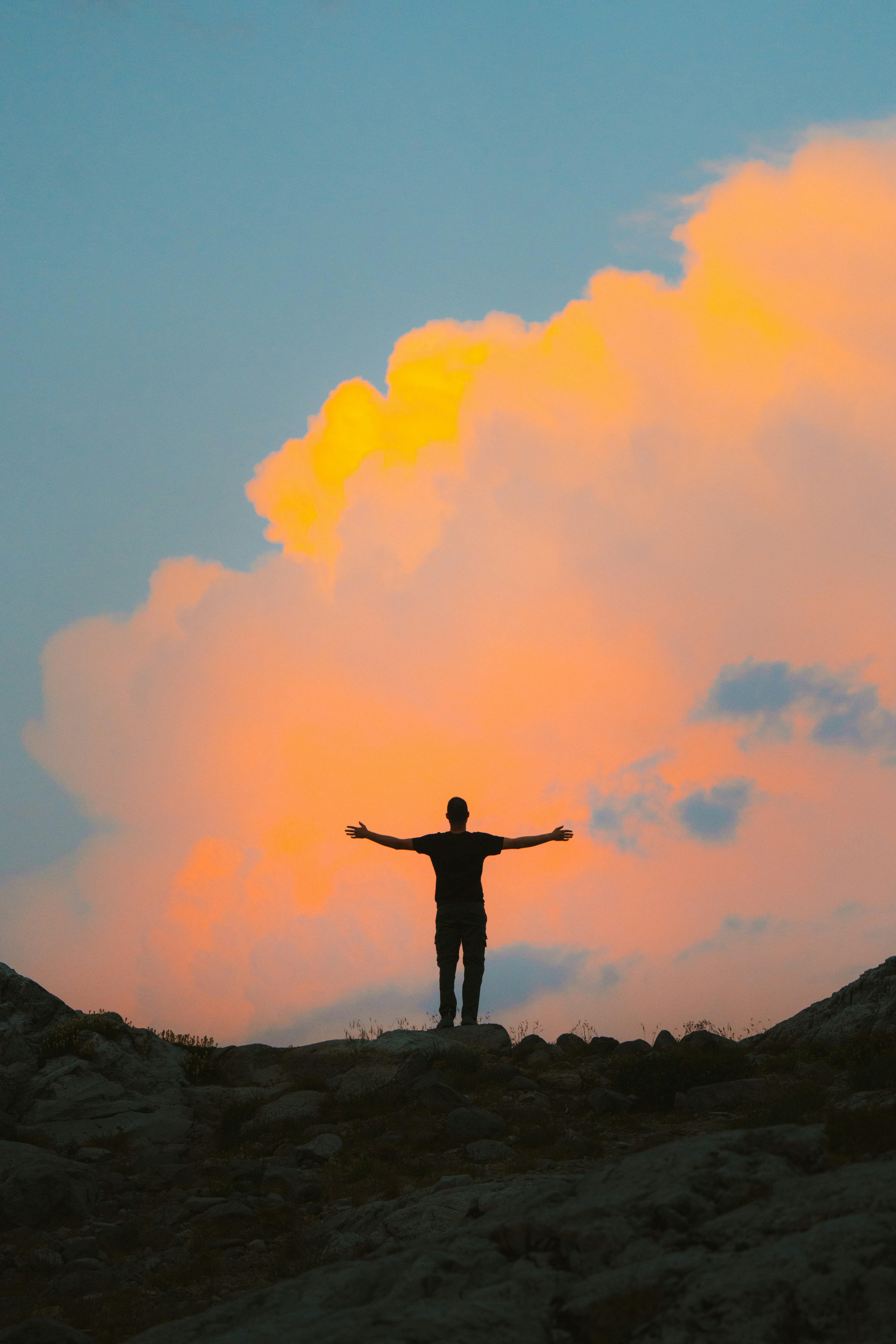 Silhouette of a person standing with arms outstretched against an orange sunset sky and clouds.