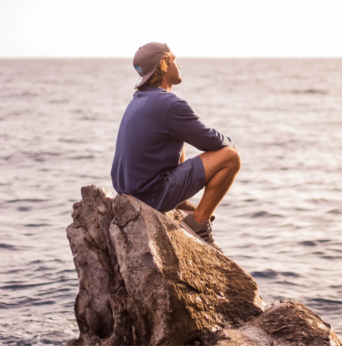 Young man wearing a cap, navy sweatshirt, shorts, and sneakers sits on a large rock by the sea, looking thoughtfully at the water.