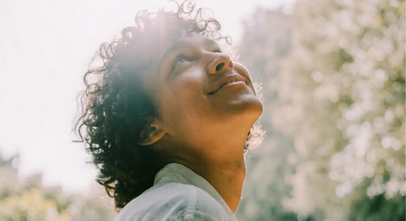 Person with curly hair smiling and looking up toward soft sunlight, a moment of relief.