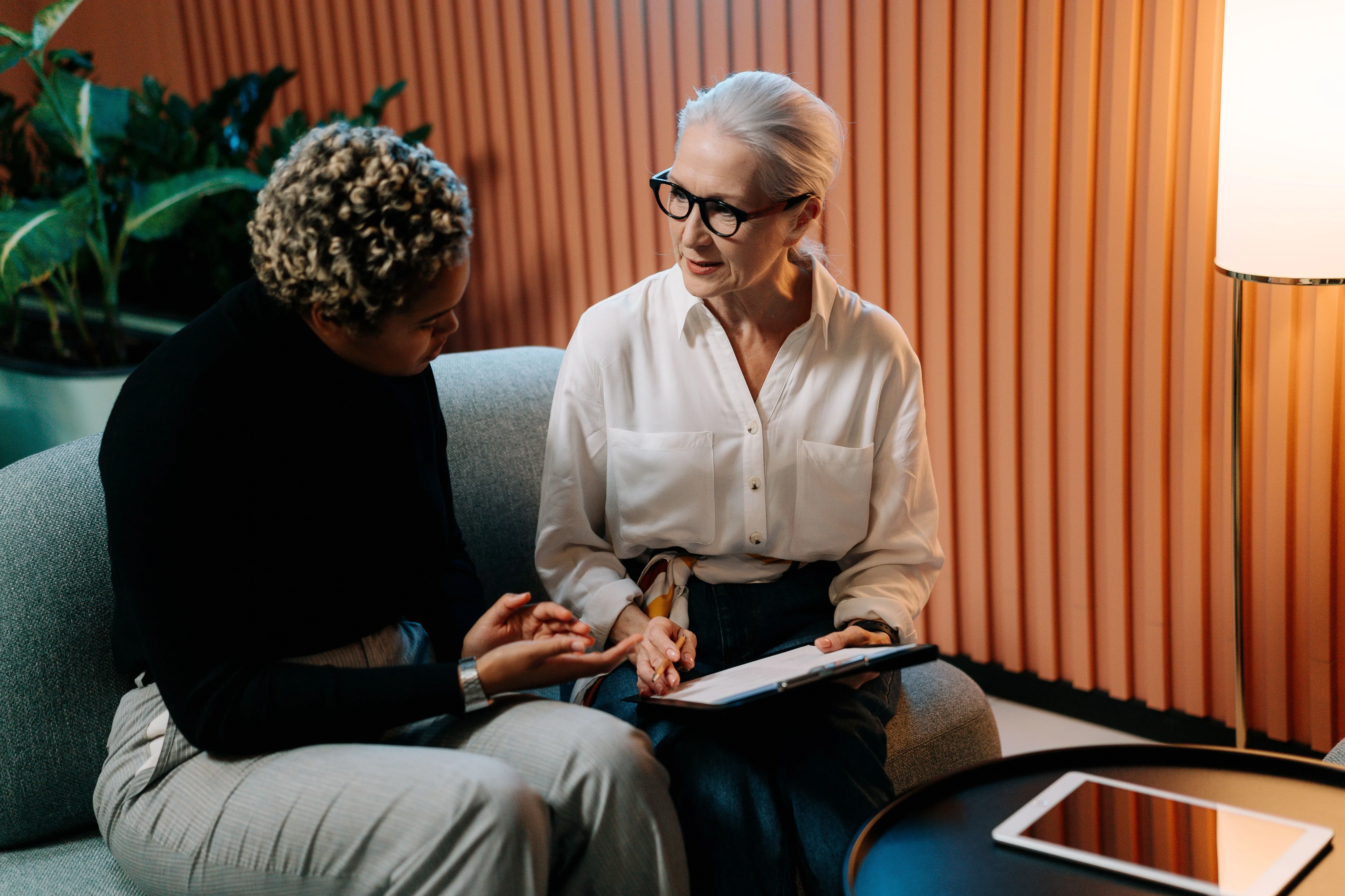 Two women sitting on a gray couch having a serious conversation, one with curly hair wearing a black top and gray pants, the other with gray hair, glasses, a white shirt, and holding a clipboard with papers.