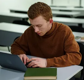Young man with short curly hair wearing a brown sweater focused on using a laptop at a desk in a classroom setting.