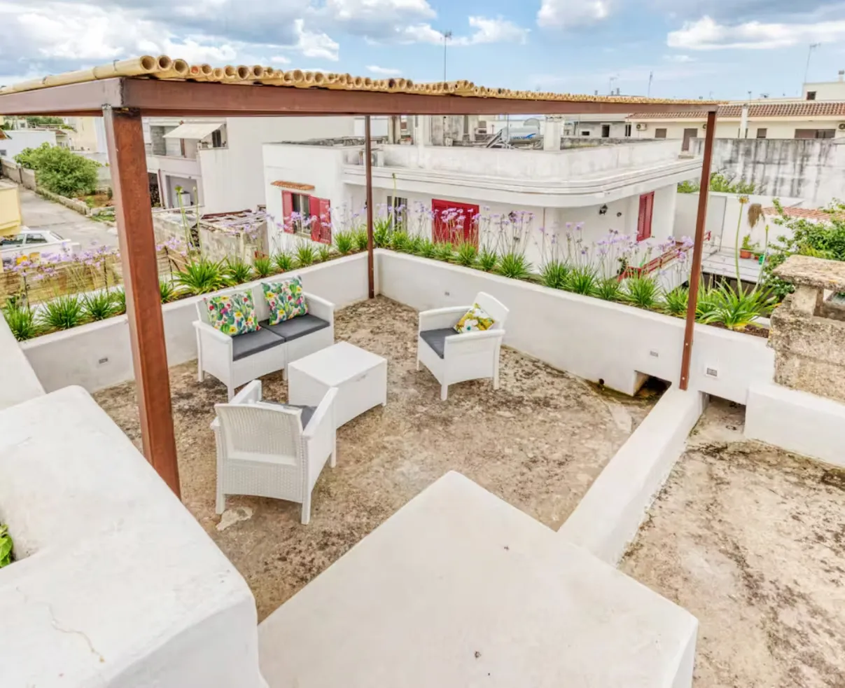 Rooftop terrace with a wooden pergola, white outdoor sofa and chairs, and flowering plants.