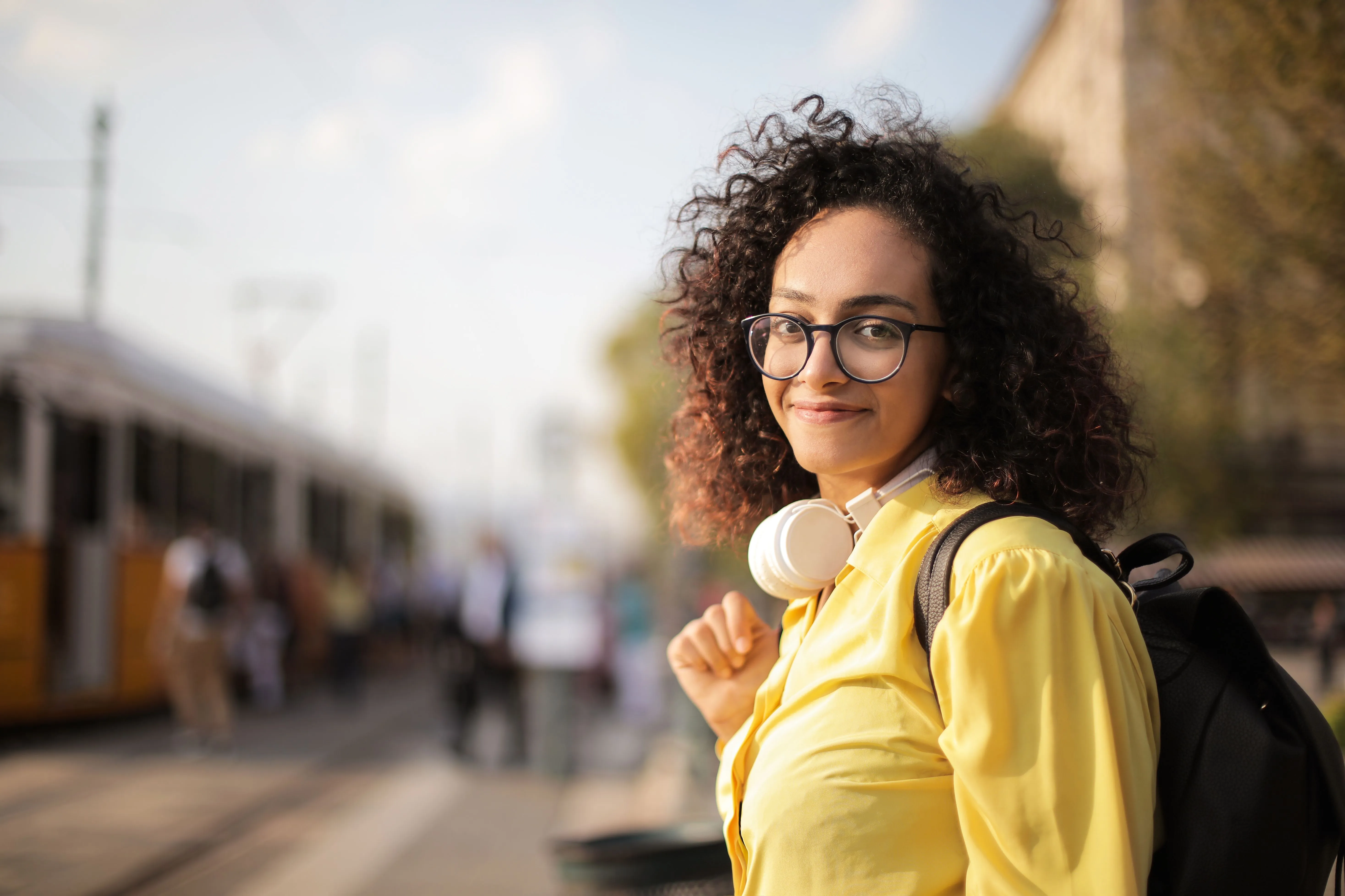 Young woman with curly hair, glasses, and a yellow shirt wearing white headphones around her neck and carrying a black backpack, standing outdoors near a tram with blurred people and trees in the background.