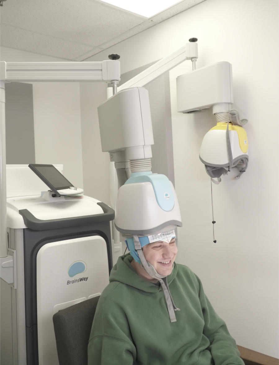 A person wearing a light blue and white BrainsWay medical helmet connected to a brain stimulation machine in a clinical setting, with another yellow helmet mounted on the wall.