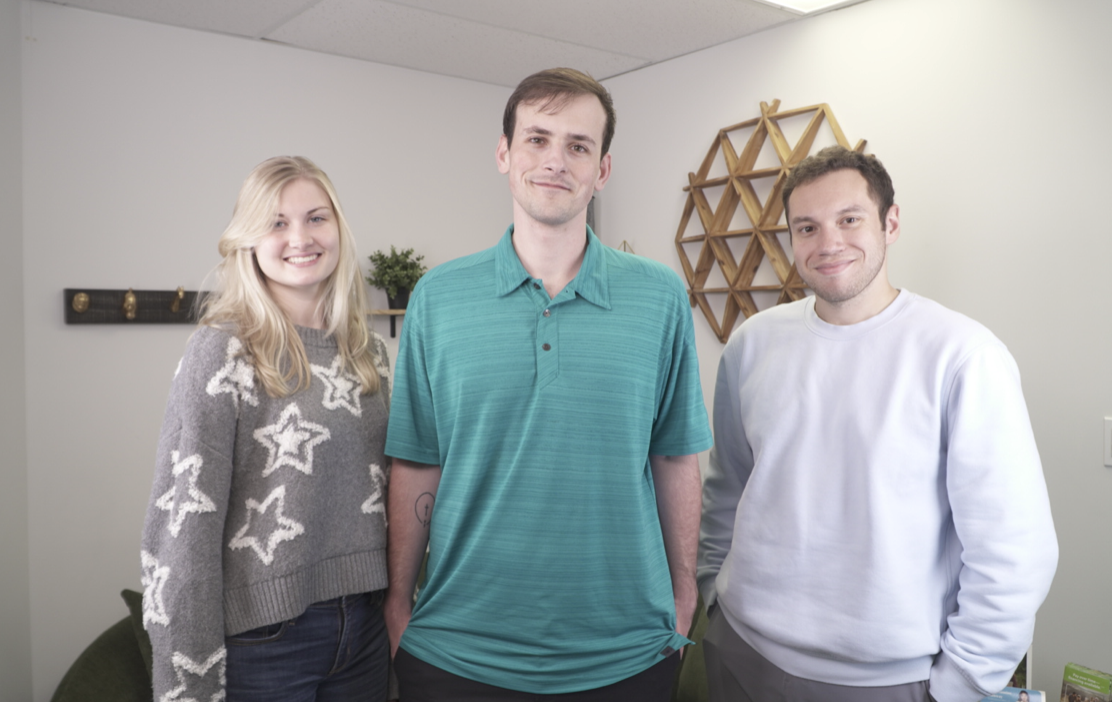Three people standing indoors, a woman on the left wearing a gray sweater with white star patterns and two men on the right, one in a teal polo shirt and the other in a white sweatshirt, smiling at the camera.