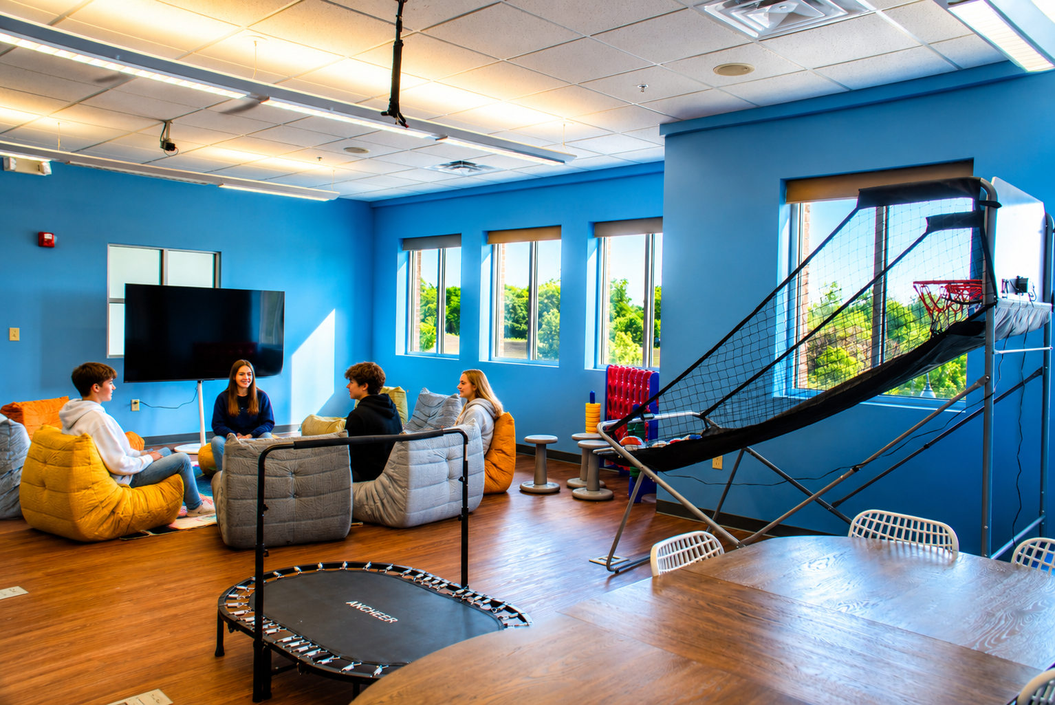Teens and a clinician talking in a bright adolescent IOP group room with bean bag chairs, a mini trampoline, and an arcade basketball game.