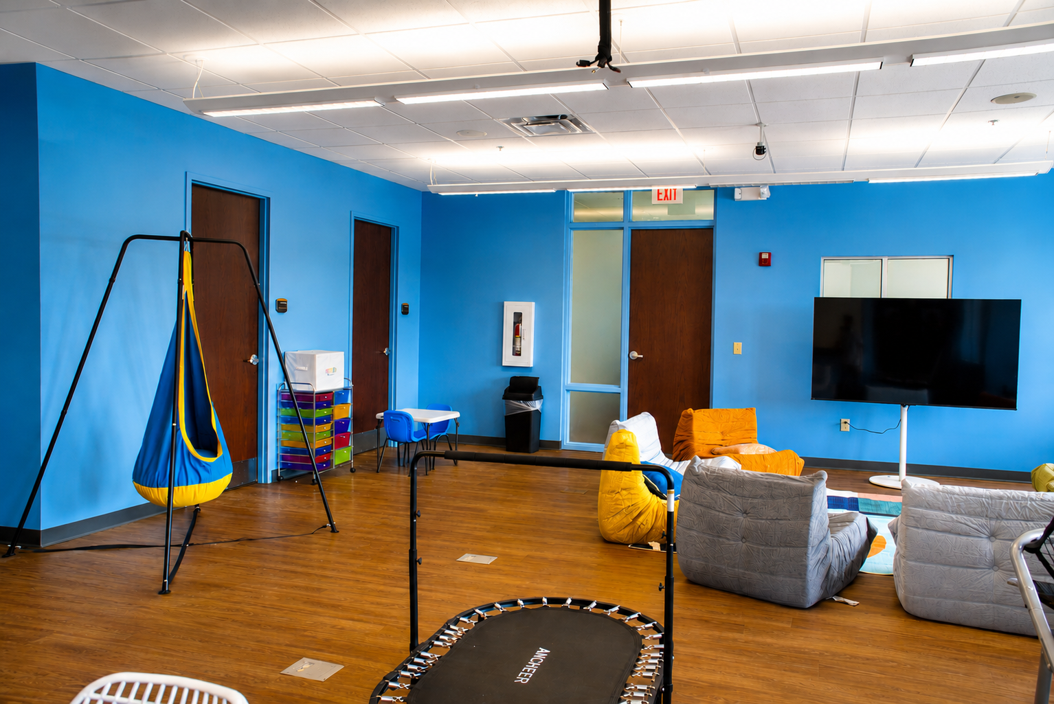 Spacious adolescent IOP therapy room at The Anxiety Center with a hanging sensory swing, mini trampoline, lounge chairs, and natural light.