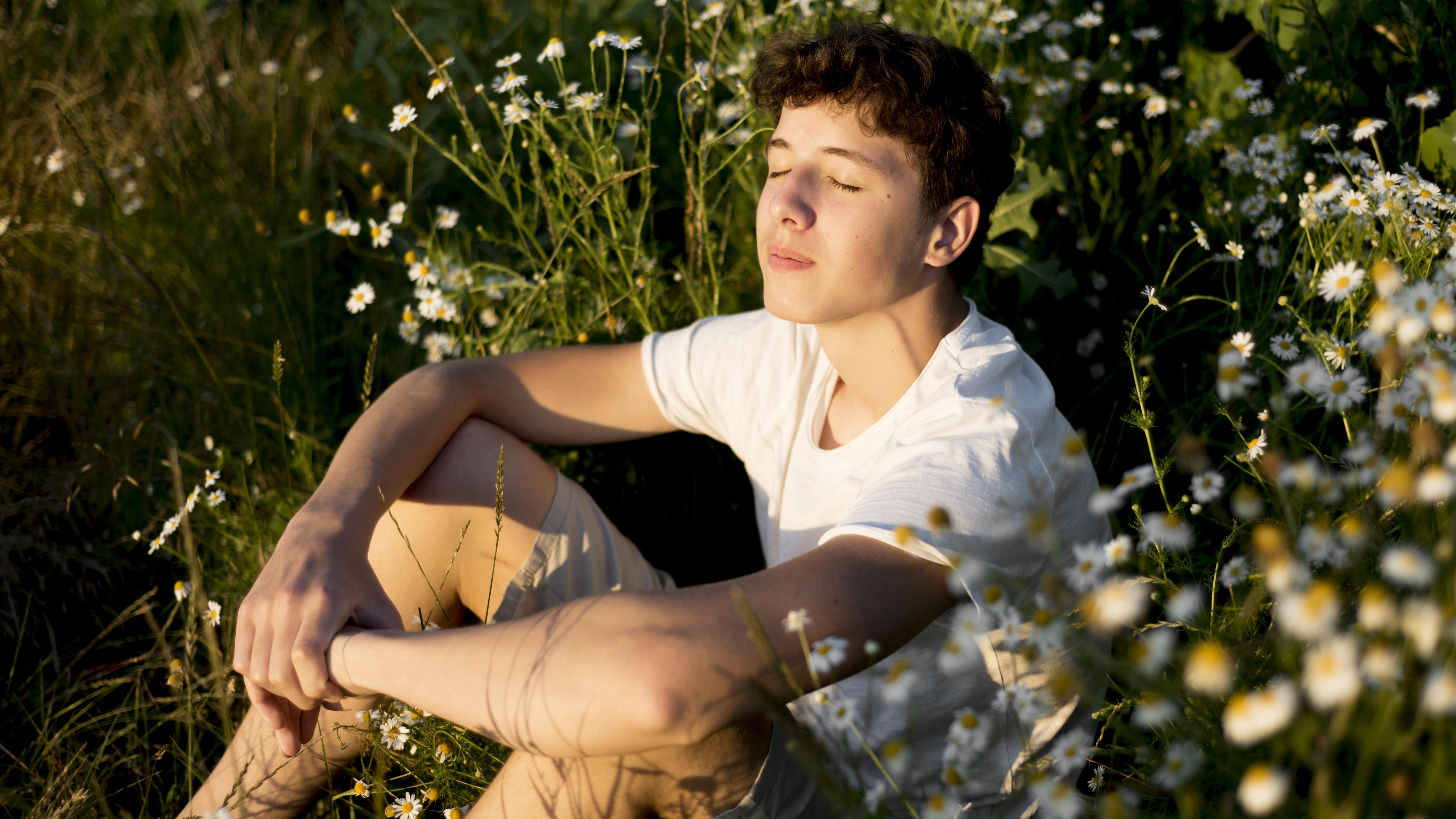 Calm teenage boy sitting with eyes closed among wild chamomile flowers in warm sunlight, a moment of peace and relief from anxiety.