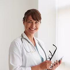 Smiling female doctor wearing a white coat and stethoscope holding eyeglasses and a tablet device, standing in a bright room.