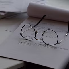 Round eyeglasses resting on an open book or document with printed text, placed on a desk with other papers nearby.