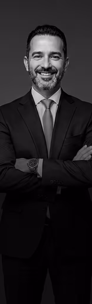 Smiling man with beard in a suit and tie, arms crossed, wearing a wristwatch, against dark background.