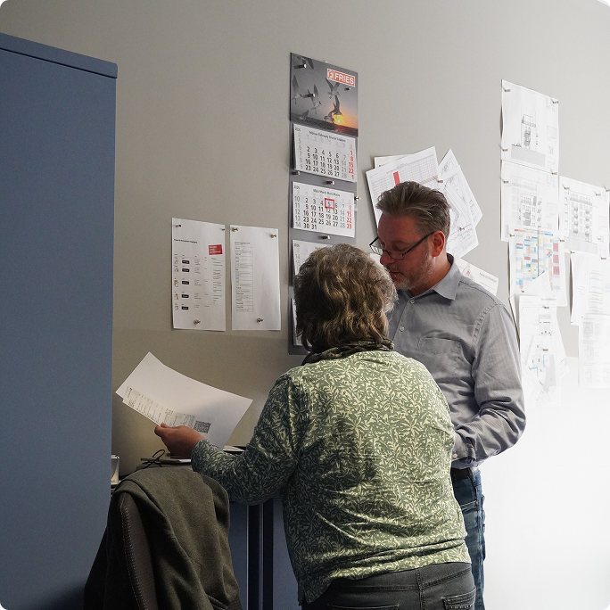 Zwei Personen im Büro vor einer Wand mit Kalendern und Plänen, eine hält ein Blatt