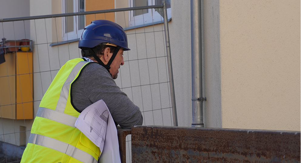 Person mit Bauhelm und Warnweste mit Bauplänen am Bauzaun vor einer Hausfassade