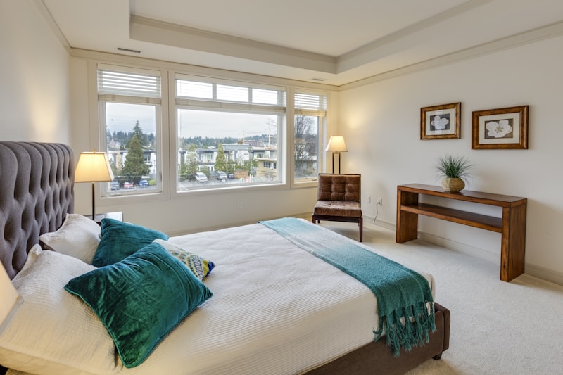 Bedroom with bed, teal pillows and throw, chair, wooden table, and window overlooking buildings.