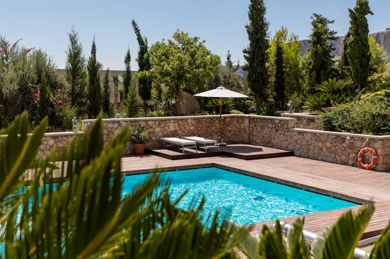 Outdoor pool with clear blue water, wooden deck, sun loungers, umbrella, surrounded by trees and plants.