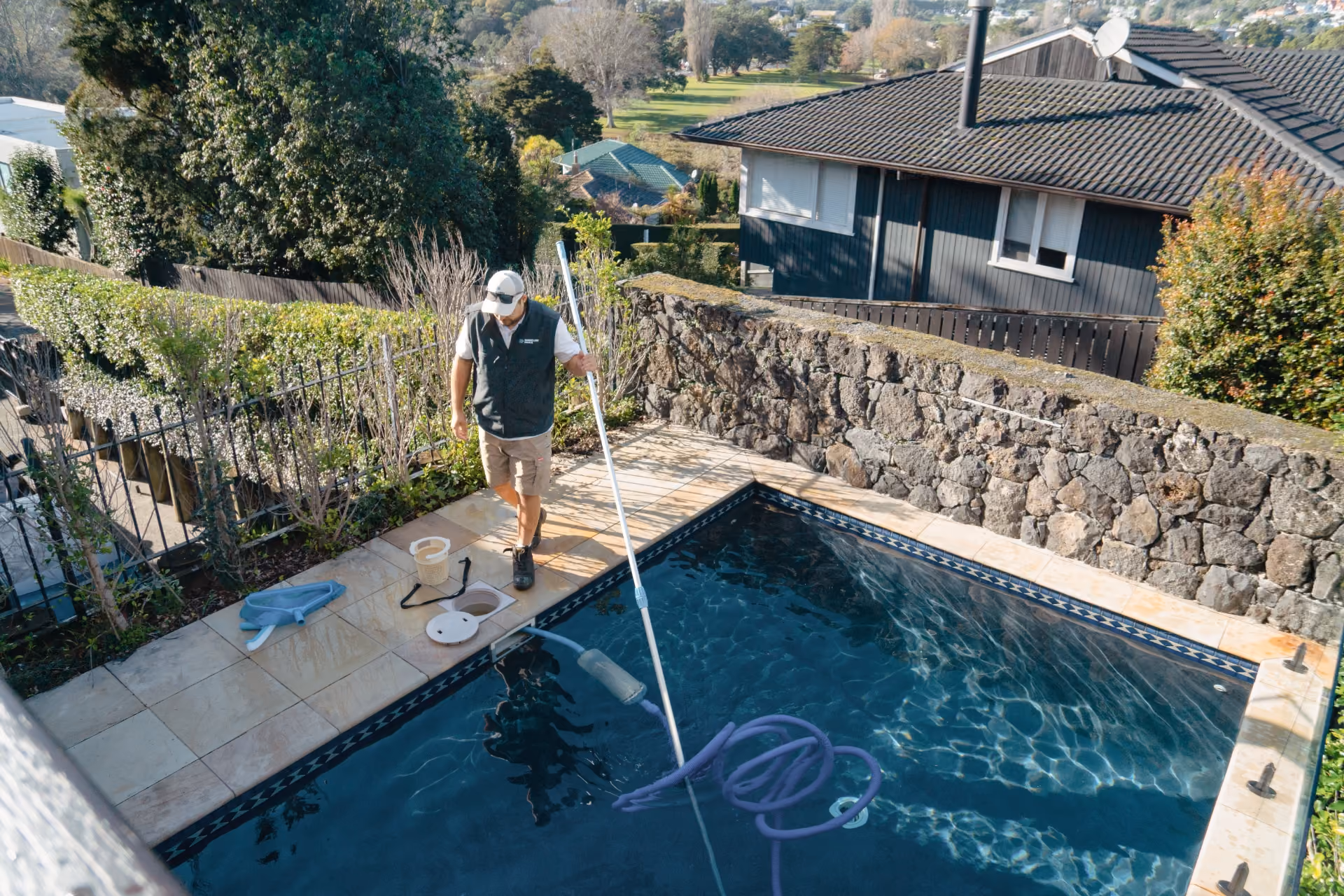 Man cleaning a residential pool with a long vacuum pole on a tiled patio near a stone wall.