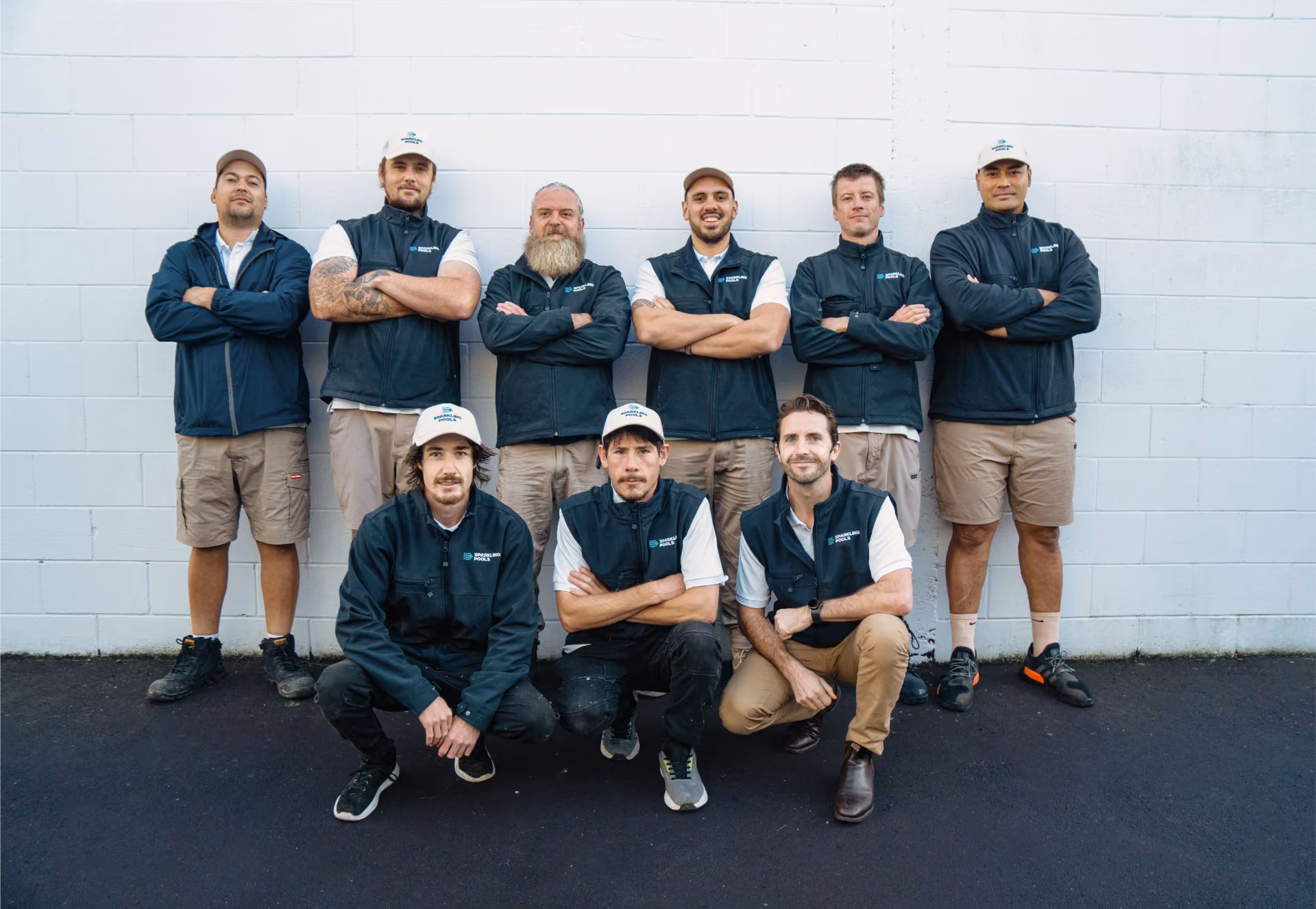 Group of nine men wearing matching outdoor workwear posing in front of a white brick wall.