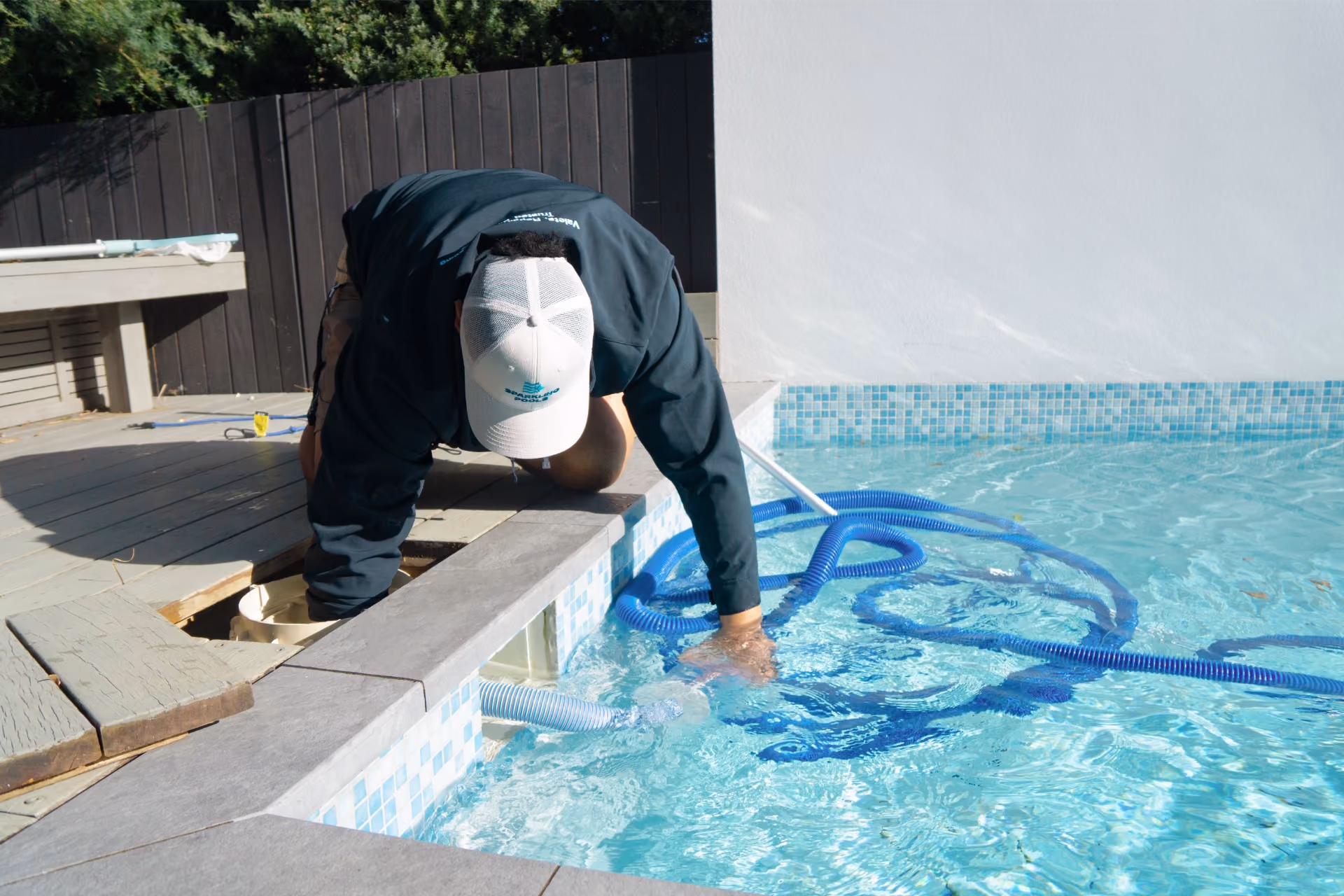 Person in cap cleaning a pool, reaching into pool water with one hand and equipment with the other.