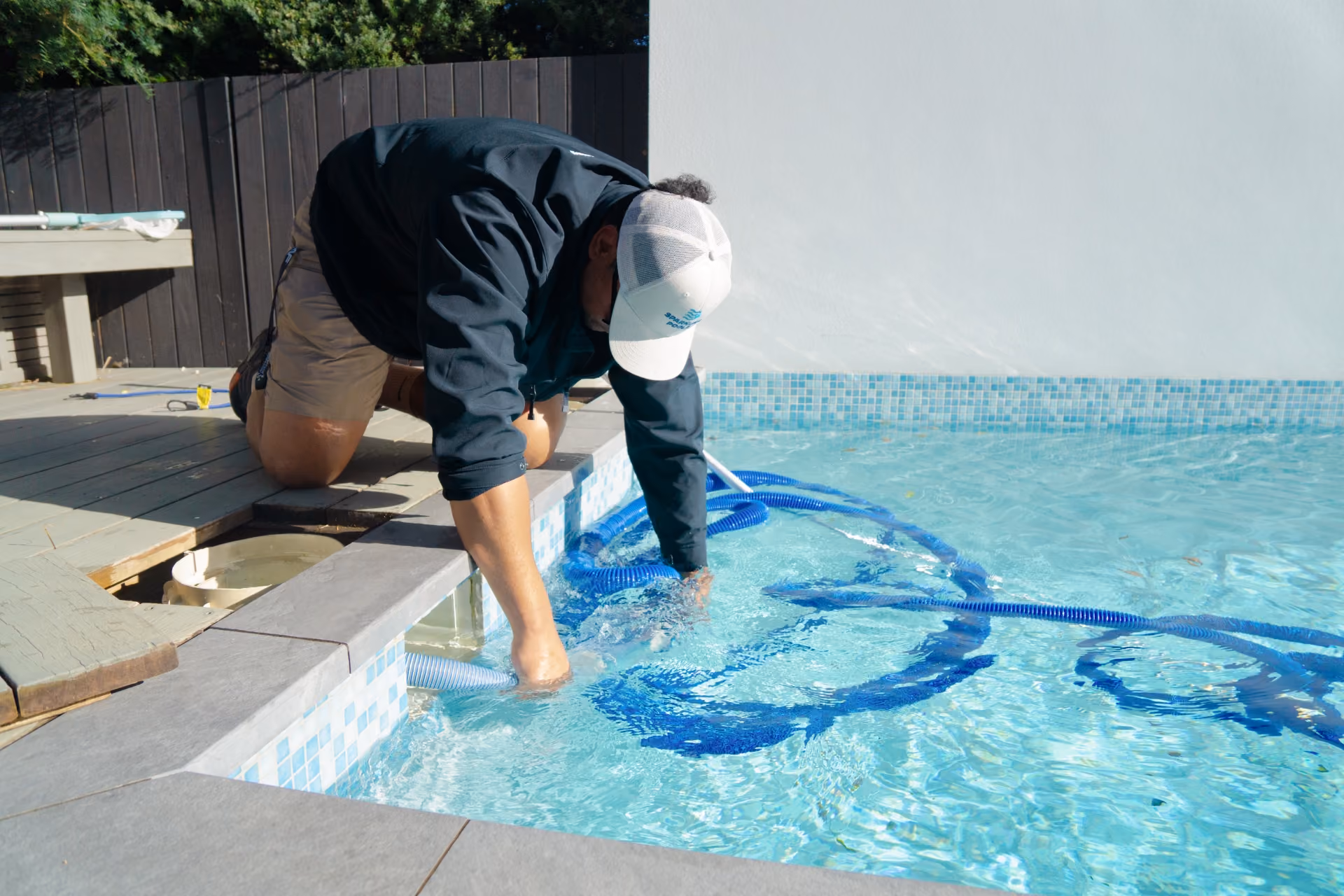 Person kneeling by the pool cleaning it using a blue hose and net skimmer.