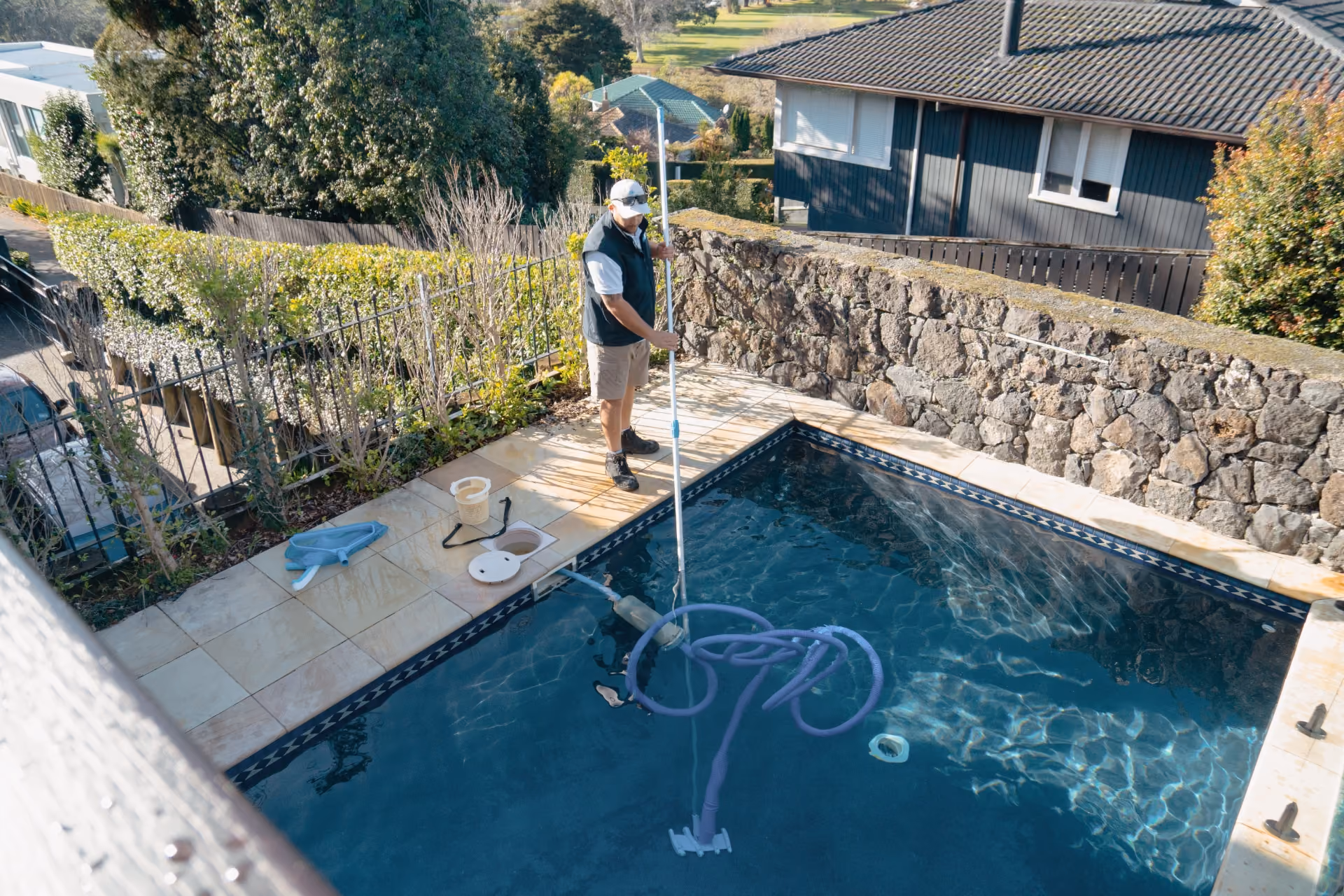 Man using a pool vacuum with blue hose to clean an outdoor swimming pool surrounded by stone walls.