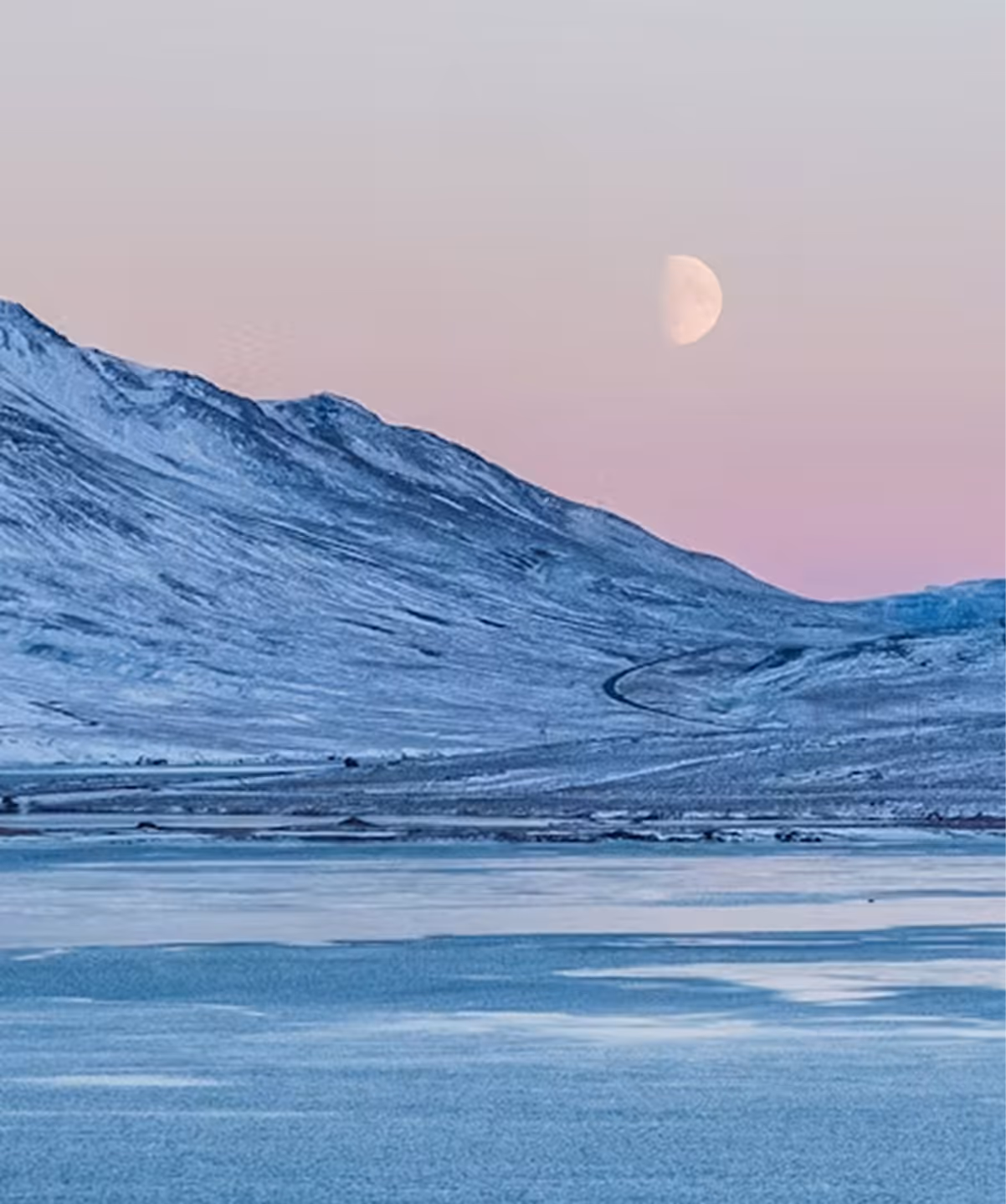Snow-covered mountains and frozen lake under a pink sky with a half moon.