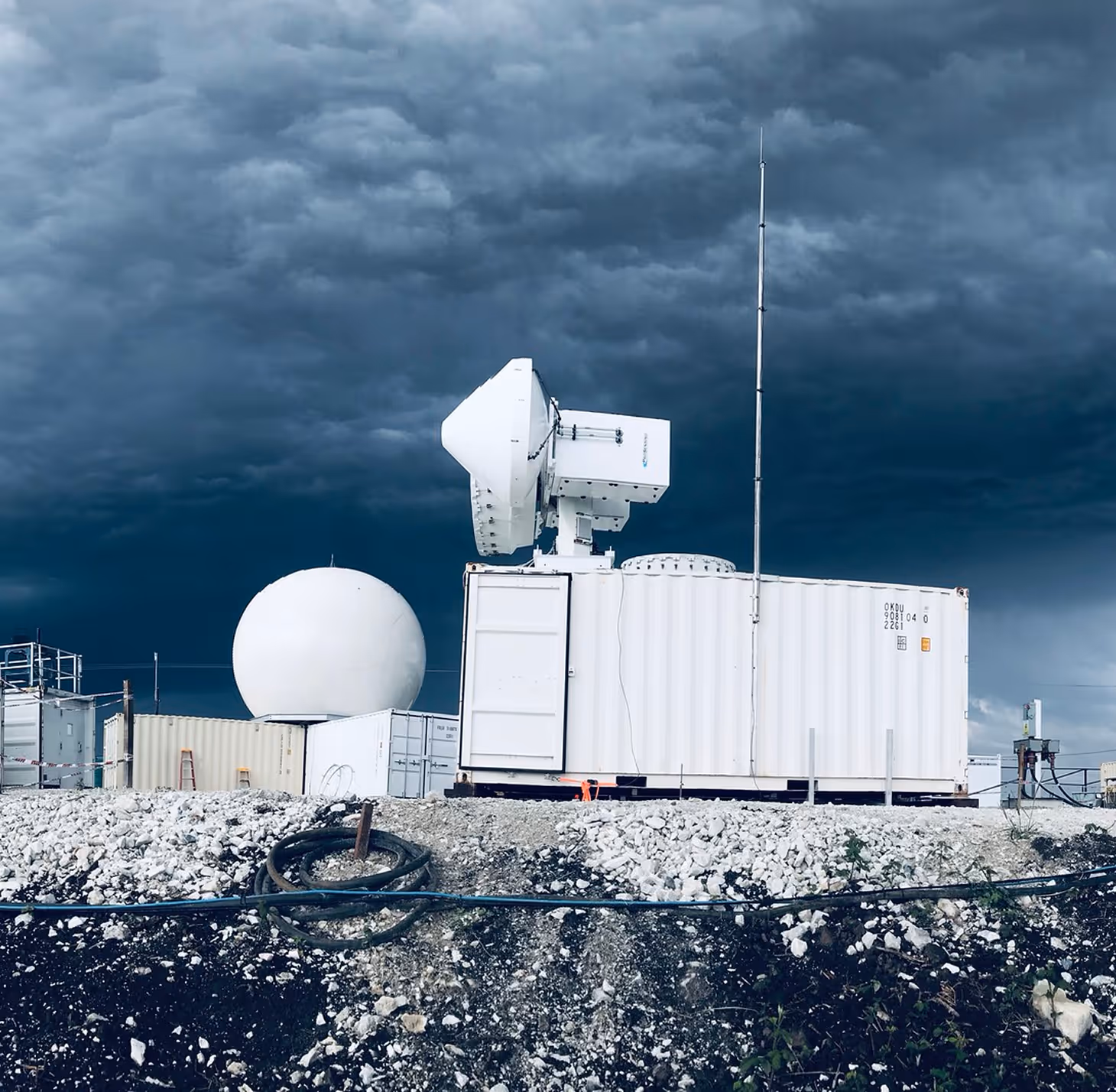 White radar and satellite communication equipment on rocky ground under dark stormy sky.