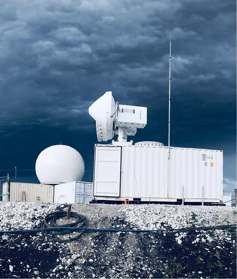 White radar installation with antenna and dome on rocky ground under dark cloudy sky.
