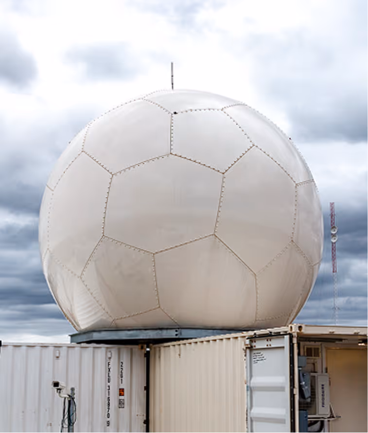 Large white geodesic radar dome atop open shipping containers under a cloudy sky.