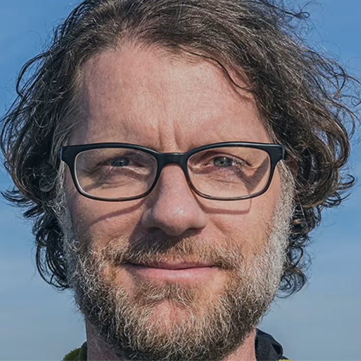 Man with curly hair, beard, and glasses looking at camera with blue sky background.