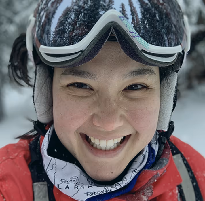 Smiling person wearing ski goggles, red jacket, and neck gaiter in a snowy outdoor setting.