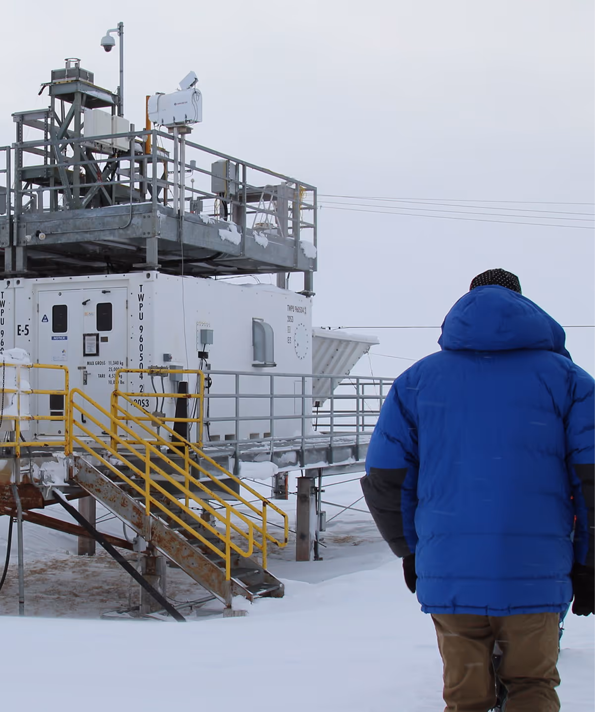 Person in blue winter jacket facing an industrial research station in a snowy landscape.