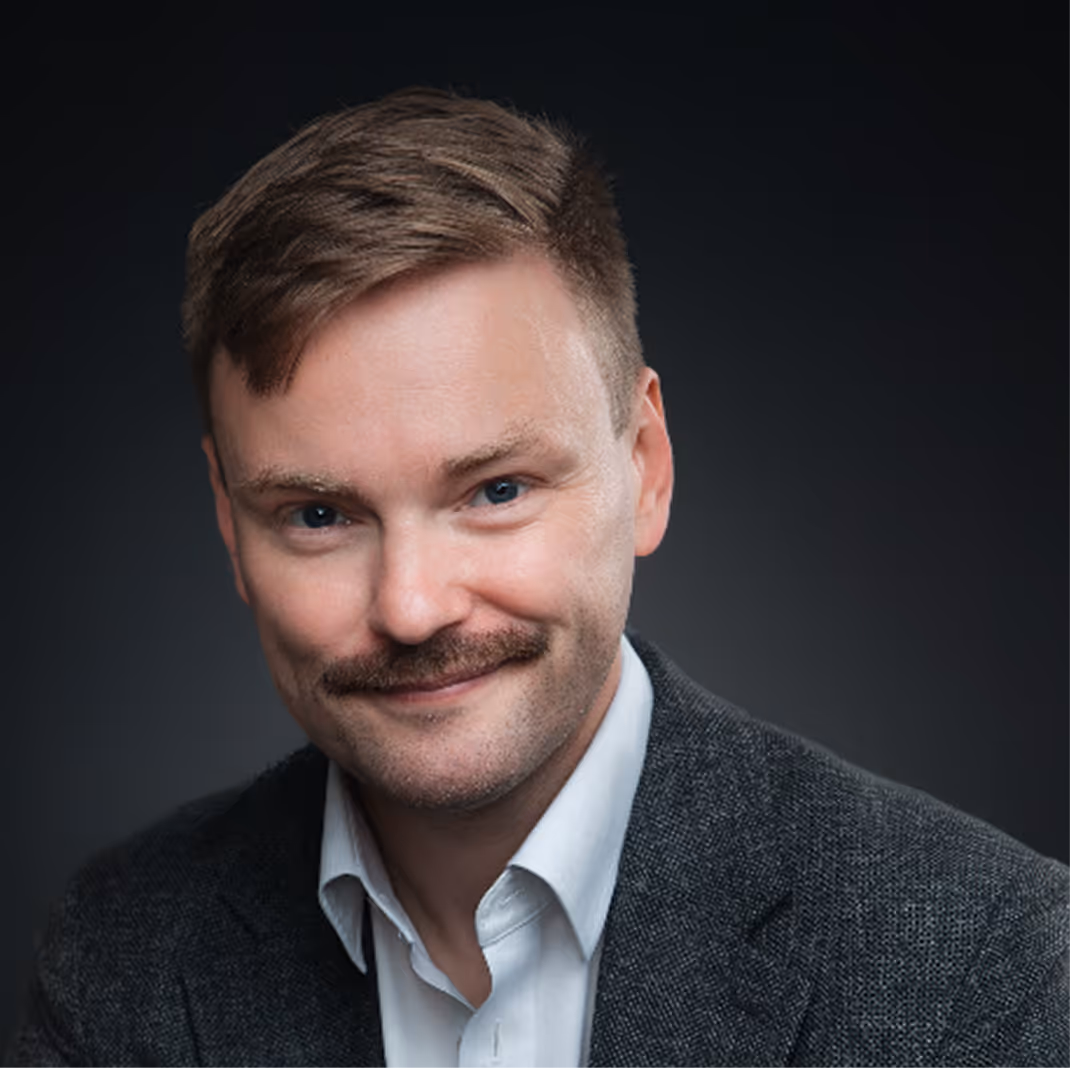 Smiling man with mustache wearing white shirt and dark gray blazer against a black background.