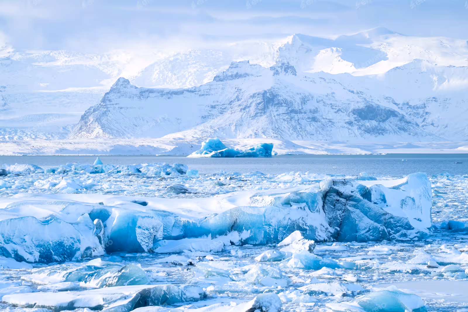 Frozen icy lake with large blue ice chunks and snow-covered mountains in the background.