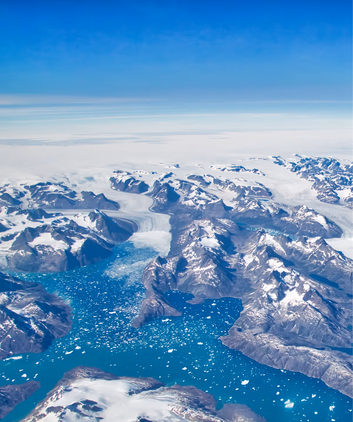 Aerial view of snow-covered mountains surrounding a blue fjord with icebergs floating on the water.