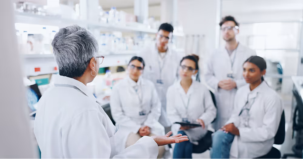 Scientist with grey hair instructing a group of attentive scientists in a laboratory.