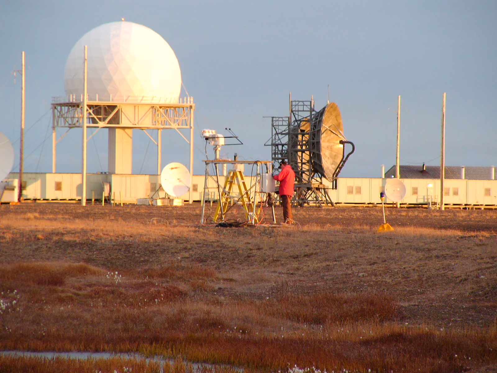 Person in red jacket working near large radar and satellite dishes on a barren field.