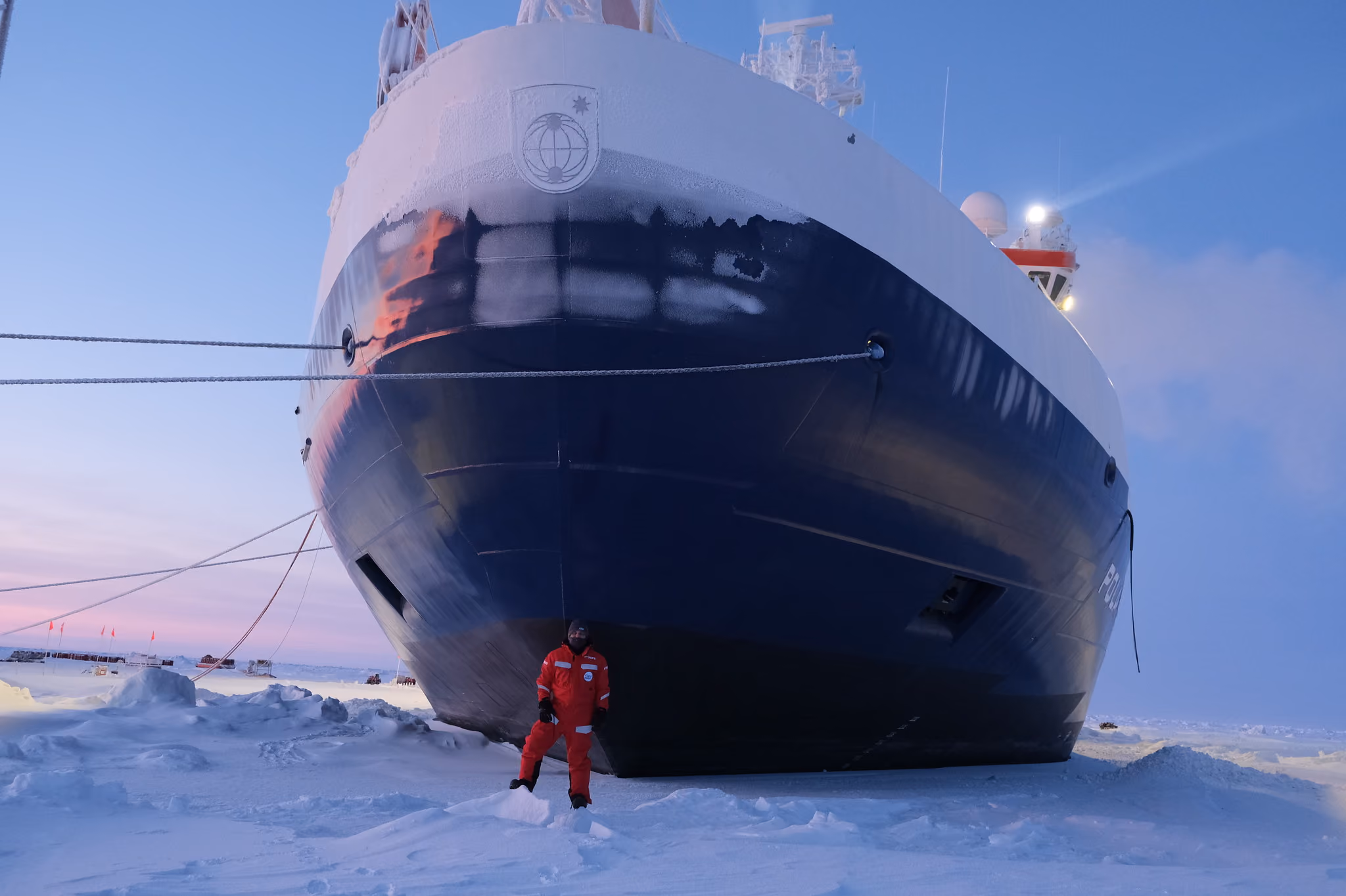 Person in red cold-weather gear standing in snow next to a large ship with icy buildup at dusk.