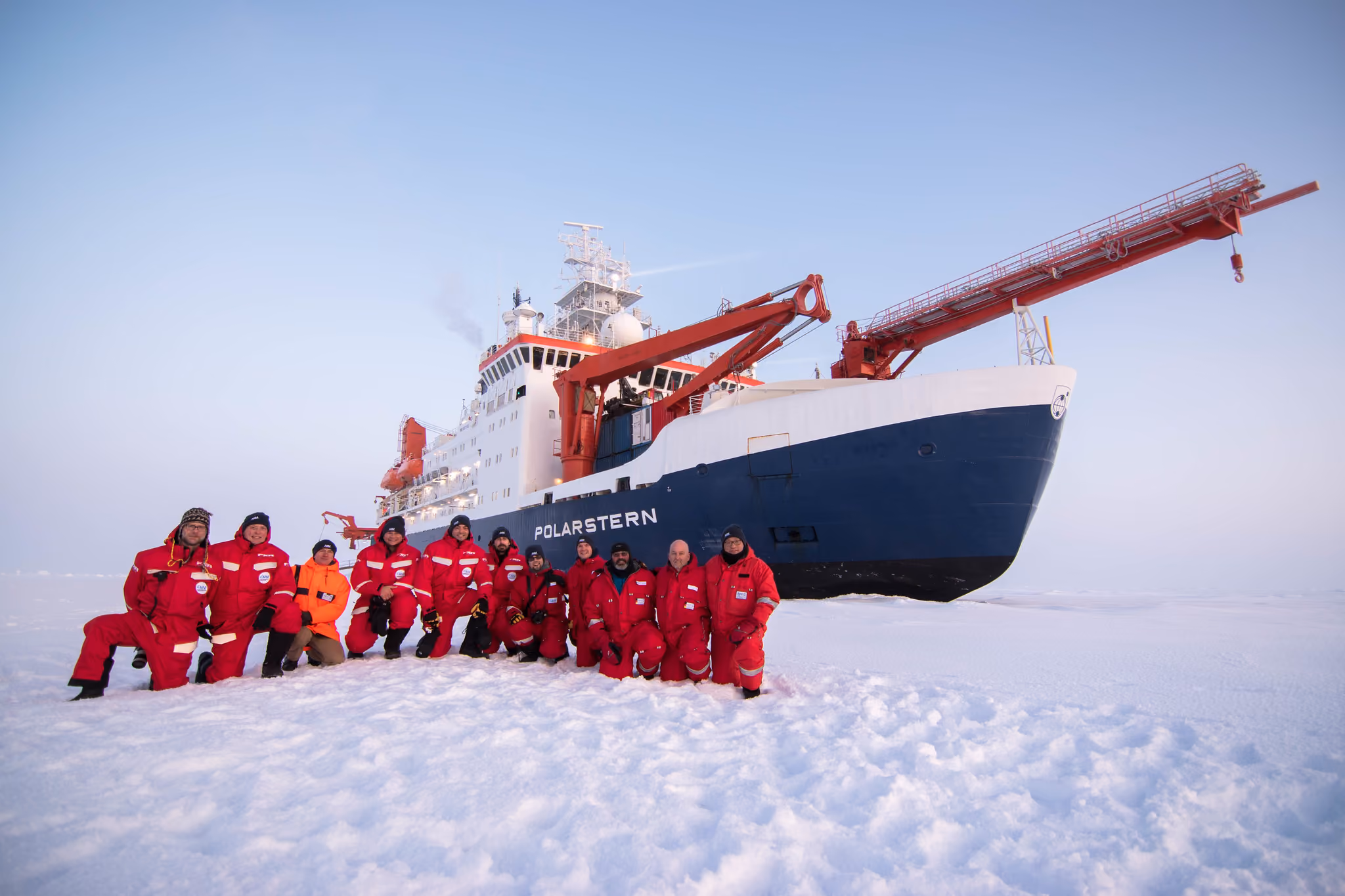 Group of people in red cold-weather gear kneeling on snow in front of the research ship Polarstern.
