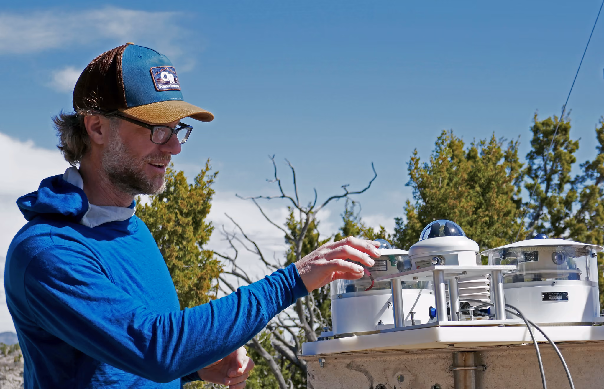 Man in blue shirt and cap adjusting a scientific instrument outdoors with trees and sky behind him.