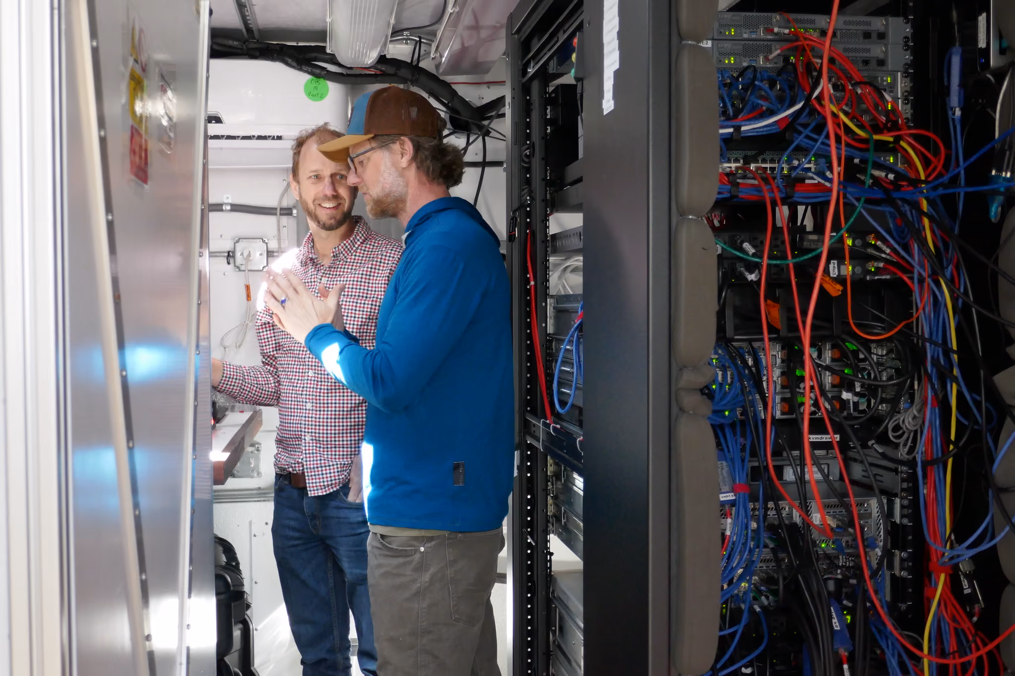 Two men discussing near a server rack with colorful network cables inside a technical room.