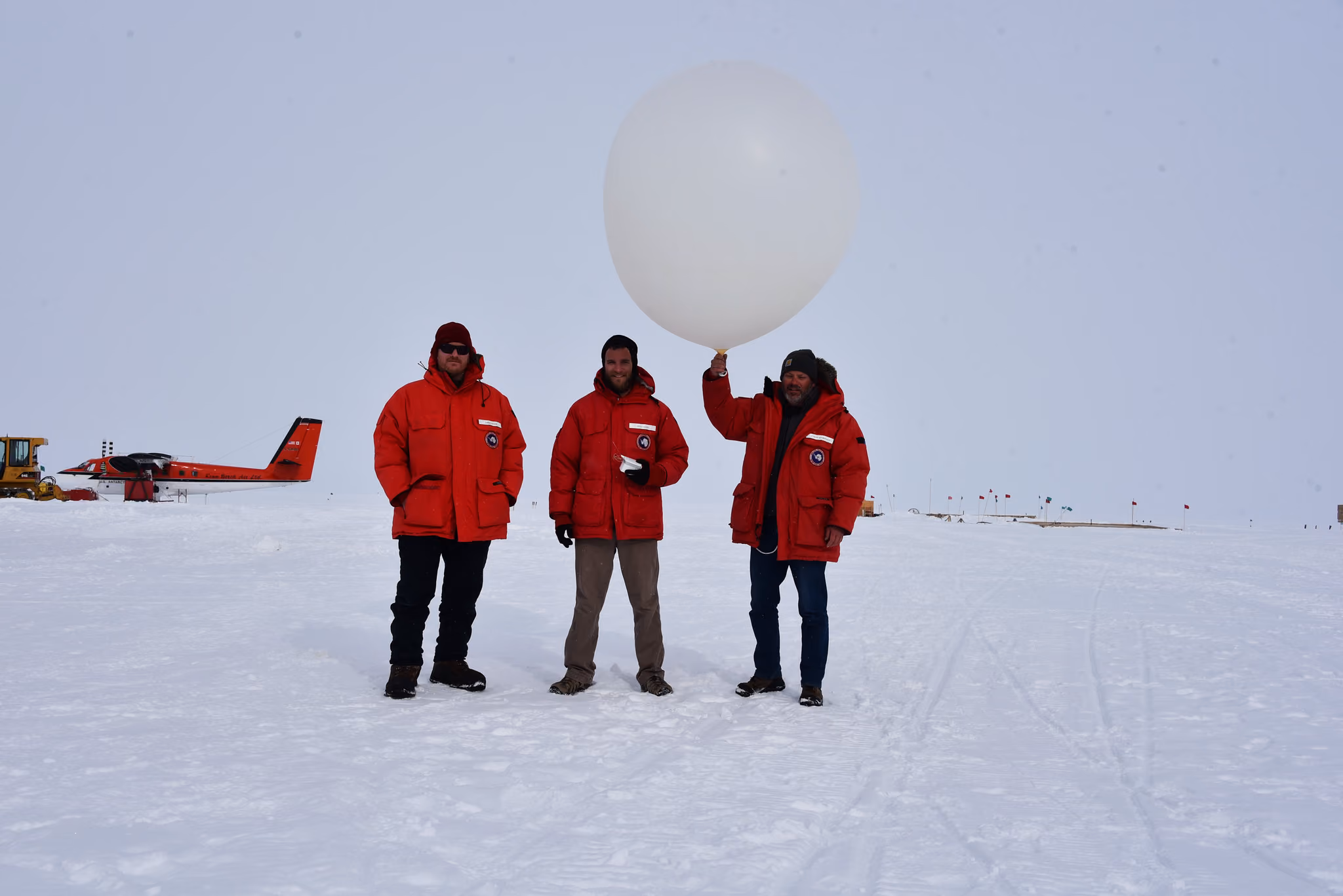 Three people in red parkas stand on snow; one holds a large white weather balloon; plane and machinery in background.