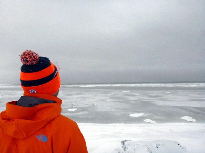 Researcher in an orange parka looking out across flat sea ice under an overcast Arctic sky.